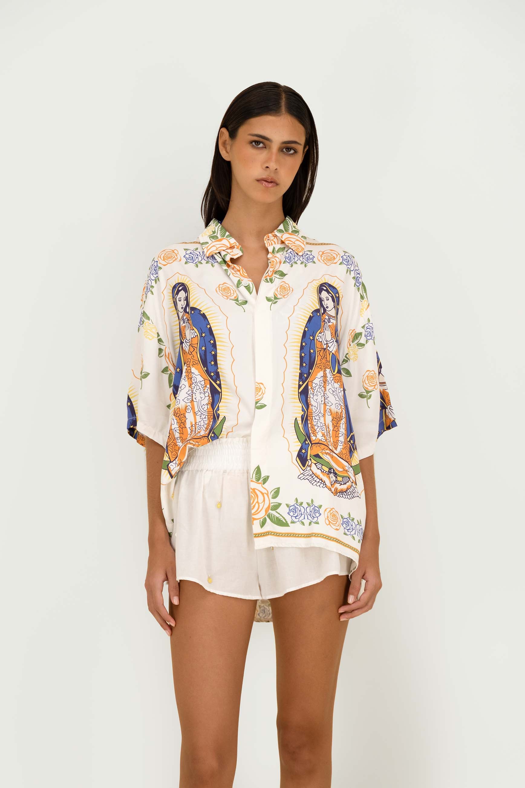 A model wearing a white short-sleeve button-up shirt and matching shorts set, featuring a colorful print of Our Lady of Guadalupe and floral motifs, against a plain white studio background.