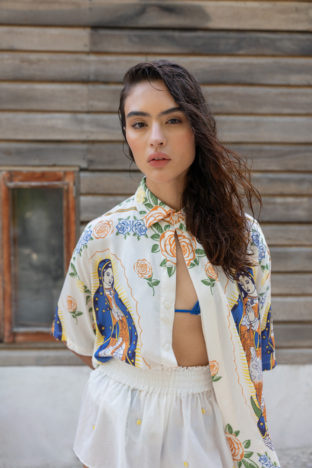 A woman wearing a white button-up shirt with colorful religious and floral prints, paired with a white skirt, standing in front of a rustic wooden wall.