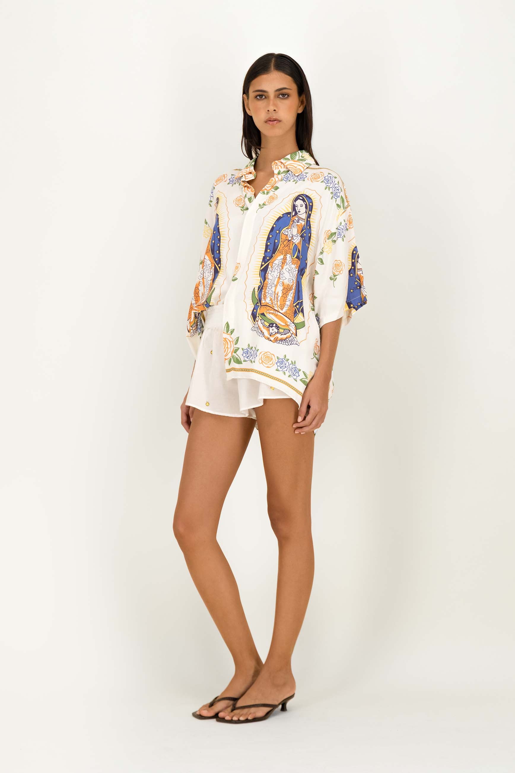 A model stands against a plain white studio background wearing a loose white button-up shirt with colorful religious-style printed figures and white shorts.