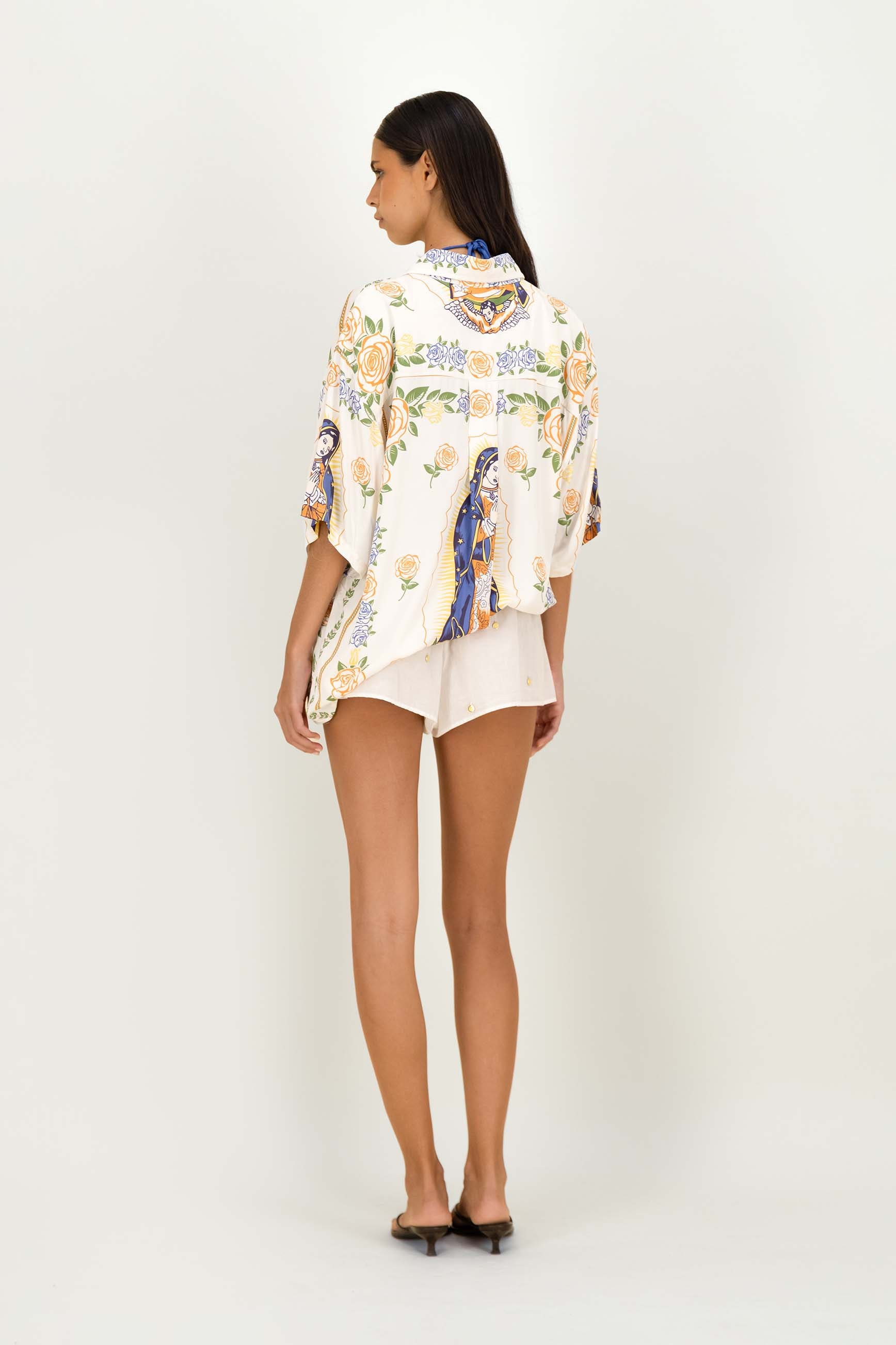 A woman is shown from the back wearing a cream-colored short-sleeve button-up shirt with a colorful floral and bird pattern, paired with white shorts and black flat sandals, against a plain white studio background.
